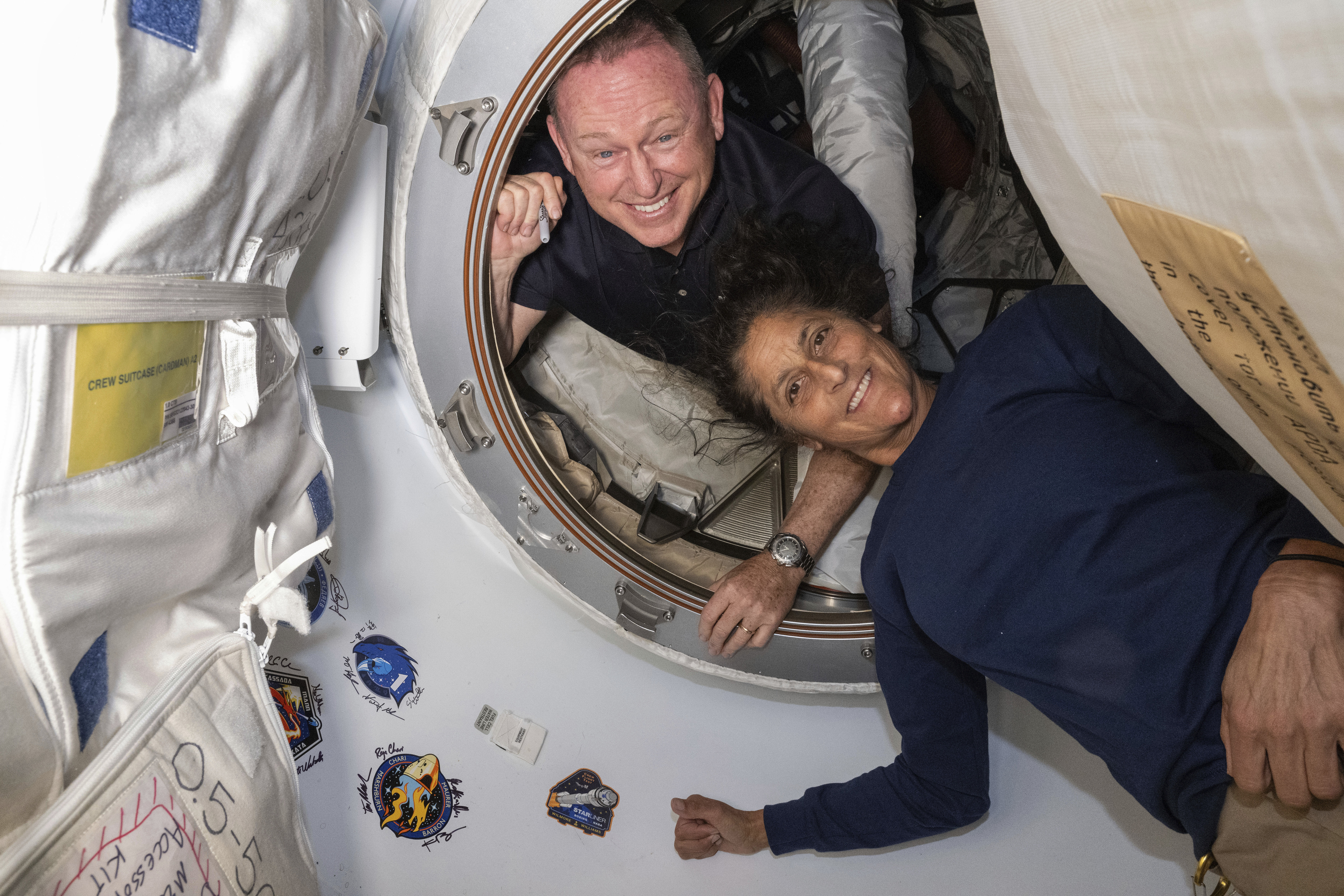 Butch Wilmore y Suni Williams, sonriendo mientras posan dentro de la Estación Espacial Internacional (ISS). Ambos se encuentran en la escotilla de la cápsula espacial, con Wilmore asomándose desde el interior y Williams acostada parcialmente fuera de la escotilla. Alrededor de ellos se pueden ver elementos del interior de la estación, como equipamiento y pegatinas con logotipos de misiones espaciales pegadas en las paredes.